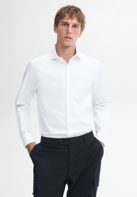 Man met lichtbruin haar, gekleed in een witte, lange mouwen blouse met knopen en donkere broek, met zijn handen in zijn zakken, staande tegen een witte achtergrond.