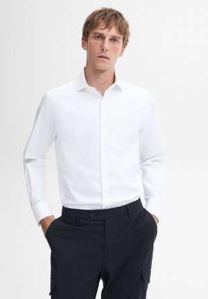 Mann mit hellbraunem Haar, der ein weißes Langarm-Hemd mit Knöpfen und dunkle Hosen trägt, mit Händen in den Taschen, stehend vor weißem Hintergrund.