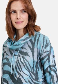 Tröja med cowl-hals, tillverkad av lätt stickad tyg i blå nyanser med ett svart abstrakt tigerstrimmönster och silverdetaljer.