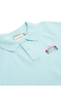 Jasnoniebieska koszulka polo wykonana z organicznej bawełny, z dwiema guzikami przy dekolcie i graficzną naszywką przedstawiającą zieloną postać w słuchawkach.