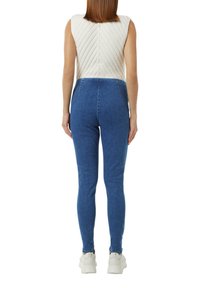 Biała bezrękawnikowa bluzka z pikowanym wzorem, zestawiona z niebieskimi legginsami dżinsowymi. Zauważalny design z wysokim stanem; model w białych sneakersach.