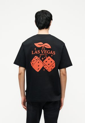 Osoba nosi čierne tričko so žiarivými oranžovými grafikami dvoch kociek, palmových listov a textom "LAS VEGAS REPLAY" na zadnej strane.