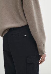 Beżowy sweter z dzianiny z prążkowanymi mankietami, w połączeniu z czarnymi spodniami cargo z bocznymi kieszeniami i małą metką na tylnej kieszeni.