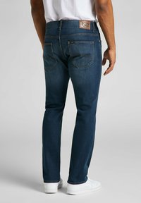 Jeans de mezclilla azul oscuro con corte recto, que presentan una etiqueta de cuero marrón en la parte trasera de la cintura y costuras distintivas en los bolsillos traseros.