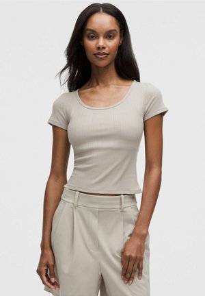 Kvinde med langt mørkt hår iført en tætsiddende kortærmet beige ribbet top og beige højtkalvede bukser, stående foran en ensfarvet baggrund.