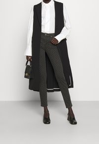 Chaleco largo negro sobre una camisa blanca, combinado con pantalones oscuros de corte ajustado y confección estampada, y zapatos negros de tacón cuadrado. Pequeño bolso negro en la mano.