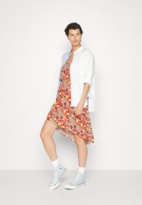 Vestido floral em vermelho, amarelo e verde com uma bainha assimétrica, combinado com uma camisa branca e ténis de cano alto azuis.