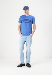 Mėlynas medvilnės marškinėlis su tamsiai mėlyna užrašu "TOMMY JEANS New York". Šviesiai mėlynos džinsinės kelnės ir juodi batai užbaigia aprangą.