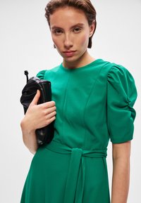 Πράσινο, κοντομάνικο φόρεμα με φουσκωτούς ώμους και φιόγκο στη μέση. Το μοντέλο κρατά μία μαύρη τσάντα με υφή. Λείος υφασμάτινος φινίρισμα.