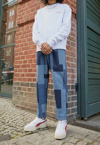 Bílý svetr s červeným textem, modré džíny s geometrickými tmavomodrými záplaty a bílé tenisky s červenými akcenty. Pozadí z cihlové zdi.
