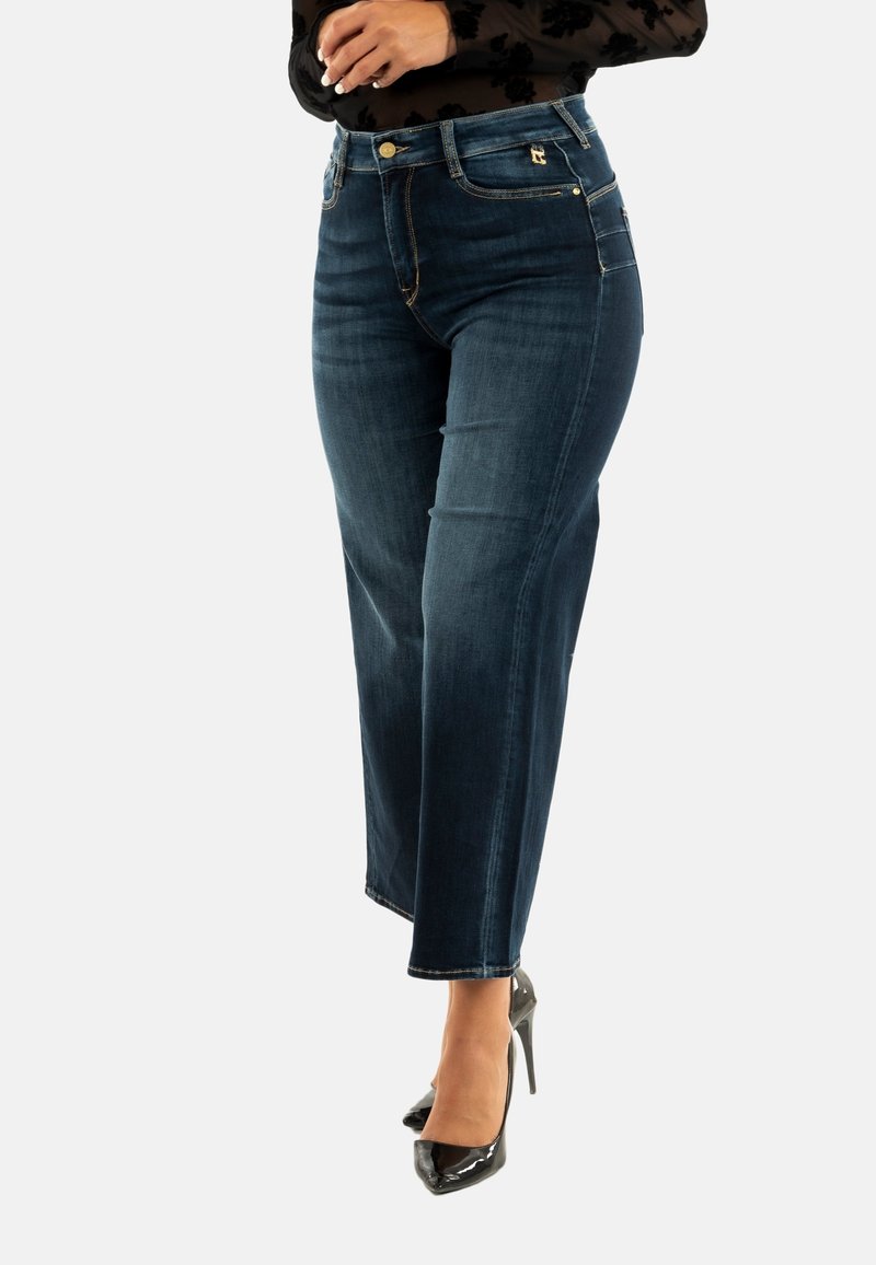 Donkerblauwe recht gesneden jeans met een licht vervaagde textuur, voorzakken en gouden accenten. Gedragen met zwarte hoge hakken.