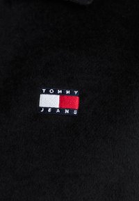 Sort fleece-stof med et rektangulært broderet logo med en blå kant, hvide, røde og blå sektioner samt teksten "TOMMY JEANS."