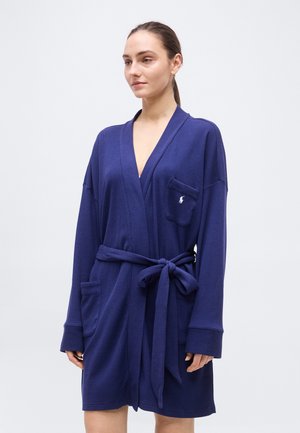 Nő térdig érő tengerészkék köntösben, amelynek derékrésznél övvel kötött, két első zsebével, és egyszínű háttér előtt áll.