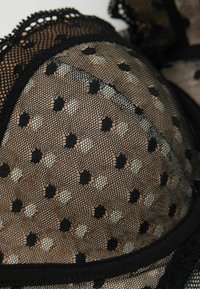 Black and beige lace bra featuring a sheer overlay with black polka dots, scalloped edges, and a black band at the bottom.
