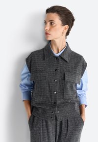 Siv zgornji del houndstooth s ovratnikom, pritisnimi gumbi in sprednjimi žepi, slojen preko svetlo modre srajce z zavihki.