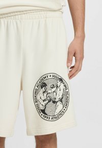 Nærbillede af cremefarvede shorts med sort cirkulær boksegraphic og teksten "Hugo Boxing Academy 1993 Athletics" på låret, ved siden af en afslappet hånd.