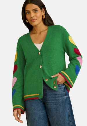 Mujer con un cárdigan verde abotonado con formas de corazones multicolor en las mangas y puños a rayas sobre vaqueros azules.