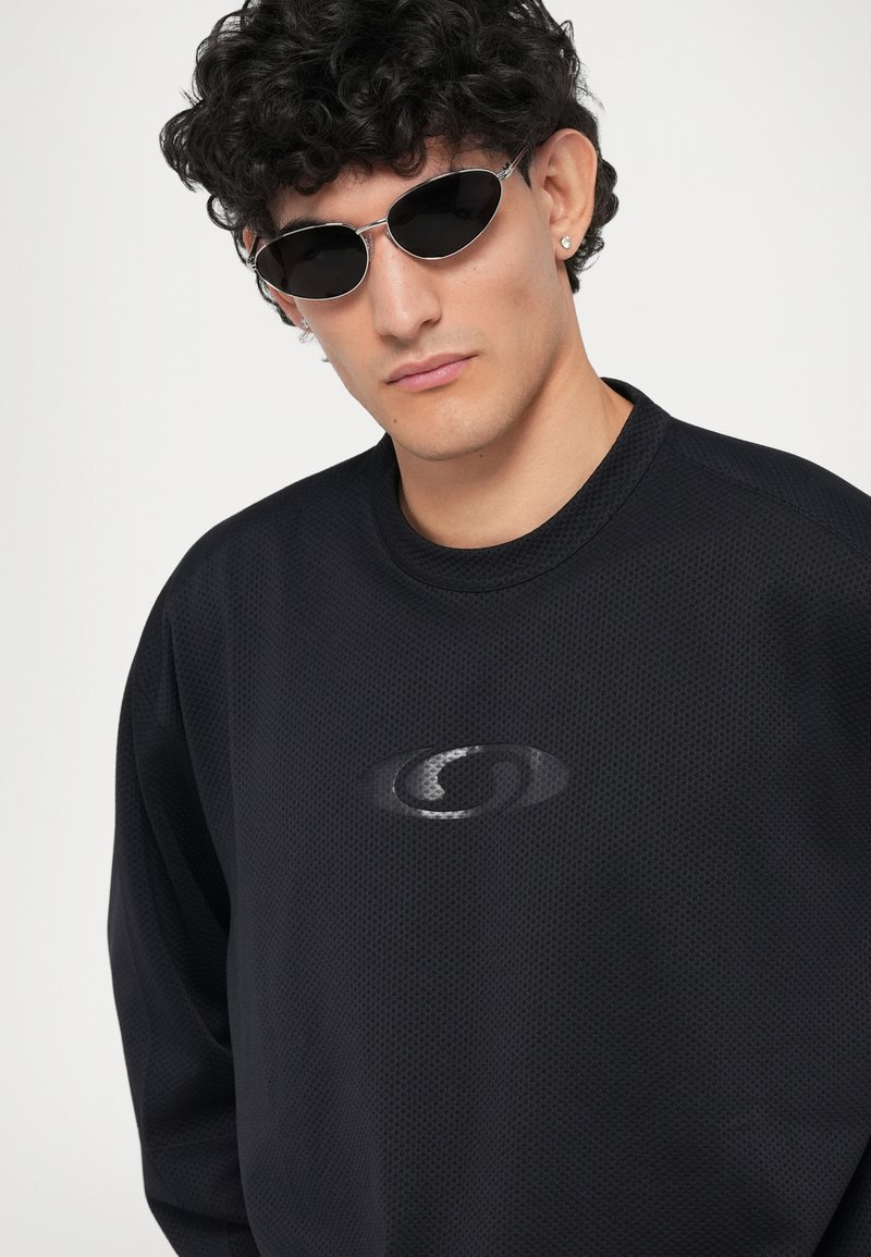 Maglietta nera a maniche lunghe con un tessuto testurizzato e un design del logo discreto sul petto. Il modello indossa occhiali da sole con montatura argentata.