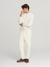Maglione bianco lavorato a maglia con polsini a coste, abbinato a pantaloni color crema e mocassini marroni da barca. Modello in piedi con aspetto pensieroso.