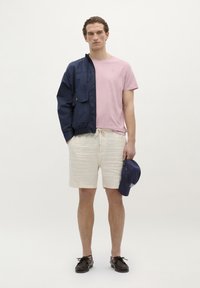 Marinblå vindjakke med lynlås foran, lyserød T-shirt med rund hals, beige linnedshorts. Bærer mørke sko og holder en marinblå kasket.