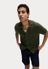 Camisa de manga corta verde oscuro con un patrón texturizado, cierre de botones en la parte frontal y cuello. Combinada con pantalones cortos de mezclilla oscura.