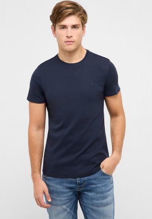 Young man with light brown hair wearing a fitted navy blue t-shirt and blue jeans, standing with one hand in pocket.