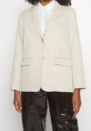 Frau trägt einen beige Blazer mit zwei vorderen Klappentaschen und Schildpattknöpfen über einem weißen Hemd mit Kragen und schwarzen glänzenden Hosen.