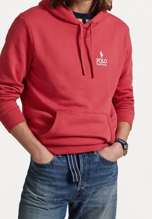 Person trägt einen roten Polo Ralph Lauren Kapuzenpullover mit Fronttasche und blaue Jeans mit gestreiftem Gürtel, Hände in den Taschen.