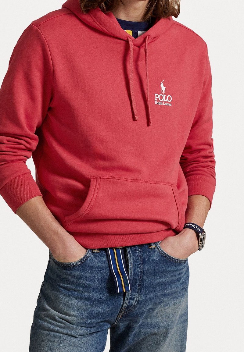Personne portant un sweat à capuche rouge Polo Ralph Lauren avec poche avant et jean bleu avec ceinture rayée, mains dans les poches.