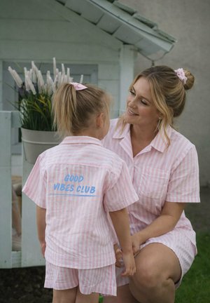 Mother and daughter in matching pink striped pajamas, daughter’s back shows "GOOD VIBES CLUB" text, outdoors near a white playhouse with flowers.