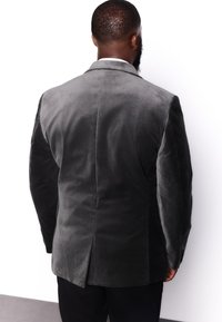 Blazer en velours gris à la texture lisse, avec une fente unique à l'arrière et une coupe cintrée. Il présente des manches longues et des épaules structurées.