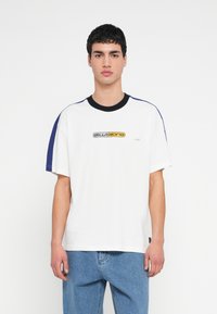 Tricou alb din bumbac cu guler negru cu nervuri, panouri laterale albastre și logo imprimat galben și negru pe față. Mâneci scurte.