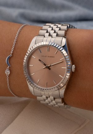Montre-bracelet en ton argent avec une lunette polie, un cadran nude et des index d'heure argentés. Accompagnée d'un bracelet délicat avec un charme en croissant.