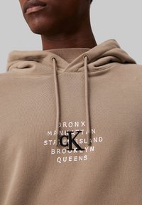 Hoodie din bumbac bej, cu buzunar frontal, șnururi și un logo imprimat care prezintă "Bronx," "Queens" și "Brooklyn" în alb și negru.