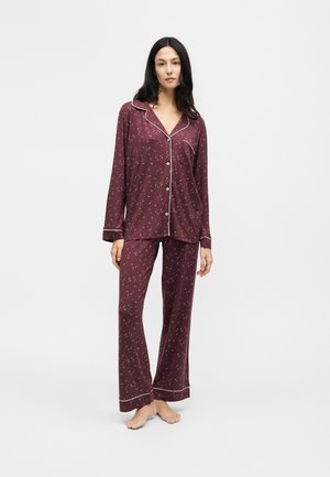 Femme debout pieds nus portant un pyjama bordeaux à manches longues boutonné avec un motif blanc subtil et des bordures blanches.