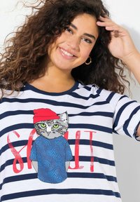 Gestreept T-shirt in wit en marineblauw met een afbeelding van een kat in een rode hoed en een blauwe shirt. Tekst "SALT" afgedrukt in het rood. Losse pasvorm, korte mouwen.