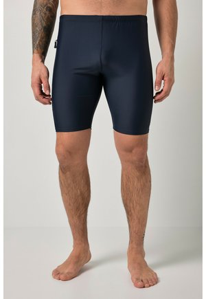 Mężczyzna bez koszulki, ubrany w przylegające ciemnogranatowe kąpielówki do kolan, stojący boso na gładkim jasnym tle.