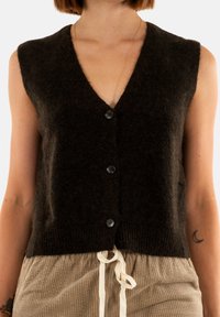 Maglione senza maniche marrone con scollatura a V, chiusura a cinque bottoni, texture morbida e orlo a costine. Indossato sopra pantaloni in velluto a coste di colore chiaro.