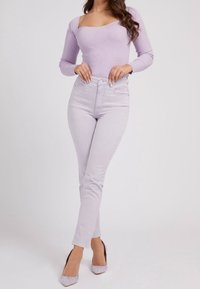 Kobieta w jasnofioletowej, prążkowanej bluzce z długim rękawem i kwadratowym dekoltem, w jasnofioletowych, wysokich spodniach oraz pasujących szpilkach.