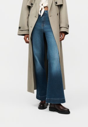 Άτομο που φοράει μπεζ μακρύ παλτό, δεμένο λευκό τοπ, μπλε τζιν ψηλόμεσο με φαρδιά πόδια και σκούρα καφέ μποτάκια αστραγάλου σε απλό φόντο.