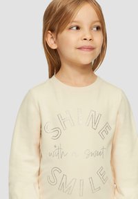 Jasno-beżowy sweter z okrągłym dekoltem, ozdobiony srebrnymi ćwiekami i napisem "ŚWIEĆ z słodkim UŚMIECHEM" oraz miękkim, teksturowanym materiałem.