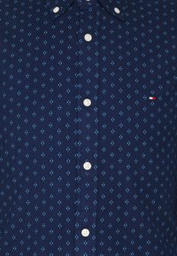 Tmavě modrá košile na zapínání s malým geometrickým vzorem v světle modré barvě. Má bílé knoflíky a malý červenobílý logo.