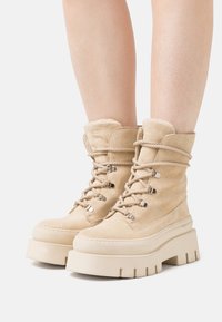 Bottes de cheville en suède beige avec une semelle plateforme haute, un laçage à l'avant et une doublure douce au niveau du col. Présente des détails en métal argenté.