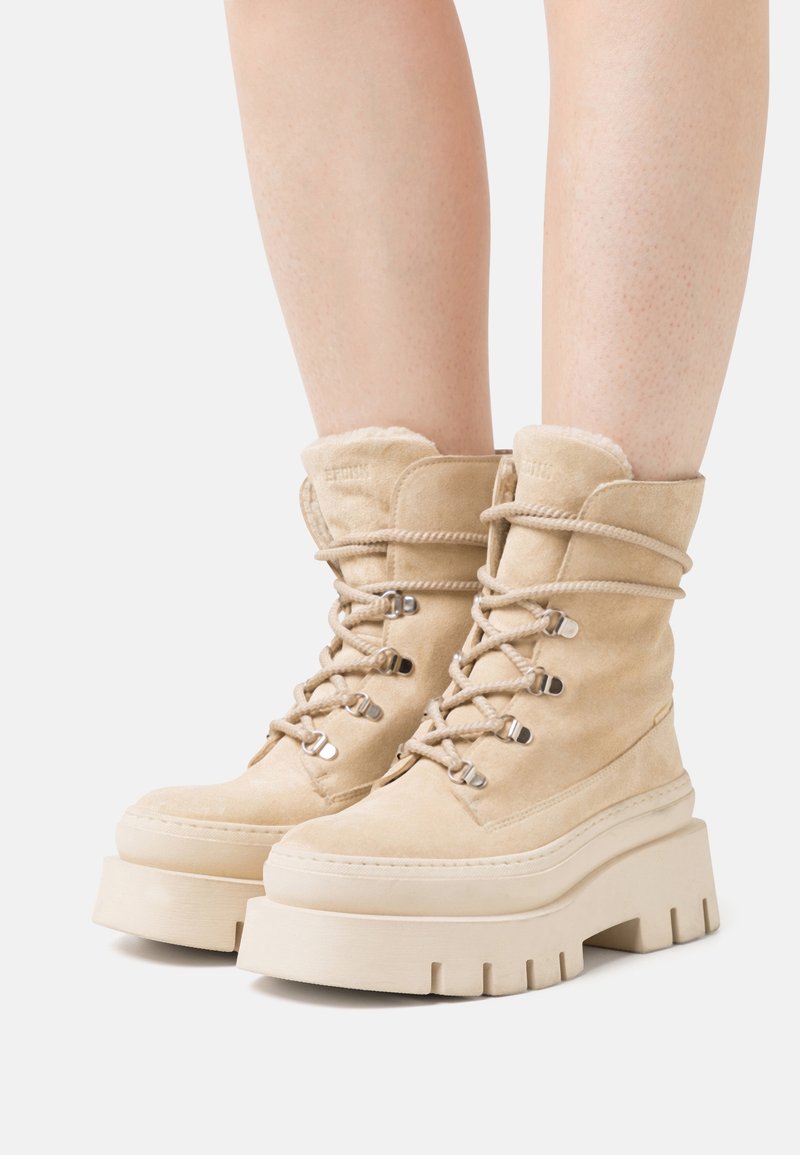 Bottes de cheville en suède beige avec une semelle plateforme haute, un laçage à l'avant et une doublure douce au niveau du col. Présente des détails en métal argenté.