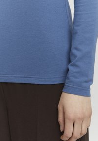 Blaues Langarmhemd aus weichem Stoff, mit rundem Ausschnitt und gesäumten Bündchen. Getragen über dunkelbraunen Hosen.