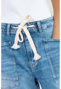 Shorts de mezclilla en azul claro con un cordón trenzado blanco que incluye borlas. Tiene grandes bolsillos laterales y un ajuste relajado.