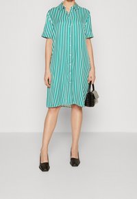 Raidallinen paitamekko teal-värissä ja valkoisena, jossa on lyhyet hihat, napitus edessä ja rento istuvuus. Mustat neliönkärkiset korkokengät ja pieni laukku mukaan.