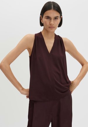 Femme aux cheveux courts et foncés portant un haut sans manches violet foncé à col en V, debout les mains sur les hanches devant un fond clair uni.