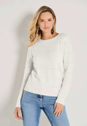 Blonde Frau, die einen weißen Strickpullover mit kleinen Löchern, blaue Jeans und große, mehrfarbige hängende Ohrringe vor einer schlichten Wand trägt.