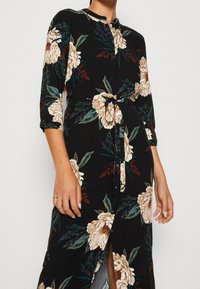Czarna sukienka w kwiaty z rękawami do trzech czwartych, z dużymi kremowymi kwiatami, zielonymi liśćmi, paskiem w talii i zapinana na guziki z przodu.